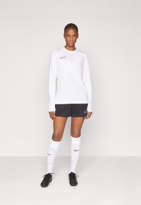 Bijela dugorukava atletska majica s crnim Nike logotipom, uparen s tamnim šortsem i bijelim čarapama s logotipom i uzorkom točkica, crnim kopačkama.