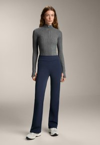 Szürke bordázott hosszú ujjú felső cipzárral, magas derekú tengerészkék nadrággal párosítva. A modell fehér tornacipőt visel. Egyszerű, modern dizájn.