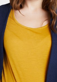 Keltainen hihaton paita pyöreällä pääntiellä, kerroksittain tekstuurinavasta kardiganin alla. Luja kangas, jossa on hienovaraisia värivaihteluita ja kiiltoa.
