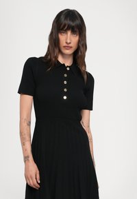Black ribbed dress with a collared neckline and short sleeves, featuring a row of gold buttons down the front and a pleated skirt.