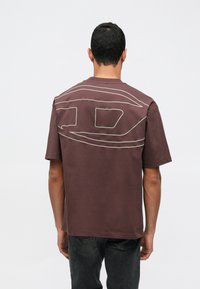 Muž otočený chrbtom, oblečený v hnedom oversized tričku s veľkým abstraktným línie dizajnom na chrbte a tmavými džínsy.