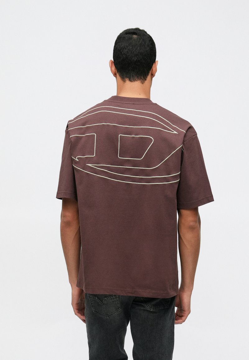 Muž otočený chrbtom, oblečený v hnedom oversized tričku s veľkým abstraktným línie dizajnom na chrbte a tmavými džínsy.