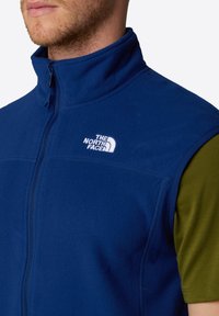 Gilet in pile blu navy con colletto alto e zip intera, dotato di logo bianco sul petto. Indossato sopra una maglietta a manica corta verde oliva. Tessuto morbido.