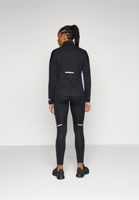 Veste de sport noire avec des accents réfléchissants et des panneaux texturés, associée à des leggings noirs avec des détails imprimés et des chaussures assorties.