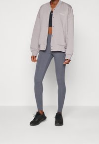 Giacca oversize grigia chiara con bottoni a pressione, abbinata a leggings grigi scuri e scarpe da ginnastica nere. Il materiale sembra morbido ed elasticizzato.
