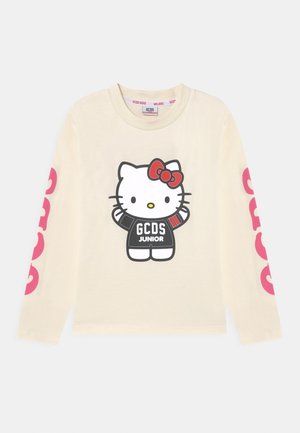 Cremefarbenes Langarmshirt mit Hello Kitty, die ein schwarzes "GCDS Junior"-Outfit und eine rote Schleife trägt, mit großem pinkfarbenem "GCDS"-Schriftzug auf beiden Ärmeln.