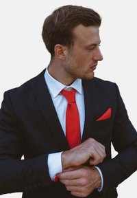 Chaqueta de traje negra sobre una camisa blanca, corbata roja y pañuelo de bolsillo. Gemelos con acentos rojos. La figura está ajustando el puño de la manga.