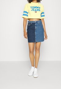 Gul kortärmad skjorta med blå ränder och text, kombinerad med en denimkjol med en tvåfärgad splittdesign. Vita sneakers fulländar looken.