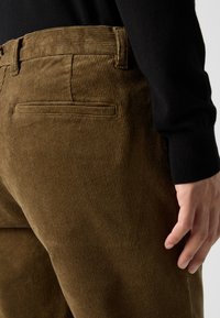 Gros plan sur une personne portant un pantalon en velours côtelé marron et un haut noir à manches longues, montrant une poche arrière et sa main droite.