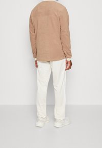 Giacca in pile color sabbia con maniche lunghe, scollo rotondo e cucitura posteriore, abbinata a pantaloni bianchi con texture e sneaker di colore chiaro.