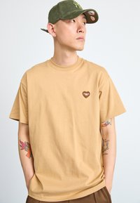 Beige cotton t-shirt with short sleeves and a round neckline. Features a brown heart-shaped logo on the left chest.