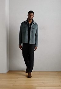 Men's casual jacket in soft gray fabric, featuring a collar, button front, and two chest pockets, paired with dark pants and brown shoes.