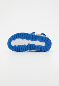 Chaussure de sport avec une semelle en caoutchouc blanche et bleue, présentant un design imprimé vibrant sur le dessus et des sangles bleues ajustables pour un maintien sécurisé.