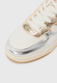 White sneaker with metallic gold and silver accents. Smooth texture with perforated details, white laces, and a light brown sole.