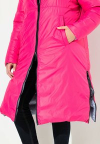 Abrigo acolchado de color rosa brillante con cremallera frontal, bolsillos laterales y forro gris. Presenta un diseño hasta la rodilla y una textura suave y brillante.