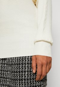 Main droite d'une personne portant un pull en maille côtelée crème reposant à côté d'une jupe en tweed à motif noir et blanc.