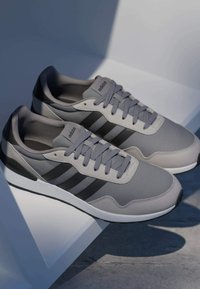 Baskets Adidas grises avec des bandes noires et des lacets gris reposant sur une surface blanche angulaire, éclairées par une lumière naturelle douce.