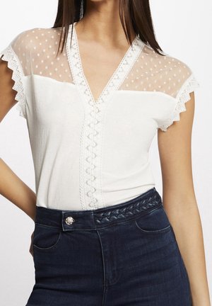 Frau trägt ein weißes V-Ausschnitt-Oberteil mit transparentem Tupfen-Spitzen-Einsatz und flatternden Ärmeln, kombiniert mit dunklen Jeans und geflochtenem Gürtel.