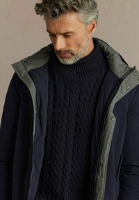 Granatowa puchówka z szarymi akcentami noszona na ciemnoniebieskim swetrze w warkocze. Kurtka posiada kaptur i zatrzaski.