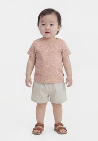 Taapero seisoo päällään pinkki kukkakuvioinen lyhythihainen paita, beiget shortsit ja ruskeat sandaalit yksinkertaista valkoista taustaa vasten.