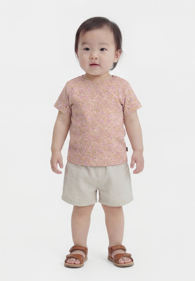 Taapero seisoo päällään pinkki kukkakuvioinen lyhythihainen paita, beiget shortsit ja ruskeat sandaalit yksinkertaista valkoista taustaa vasten.