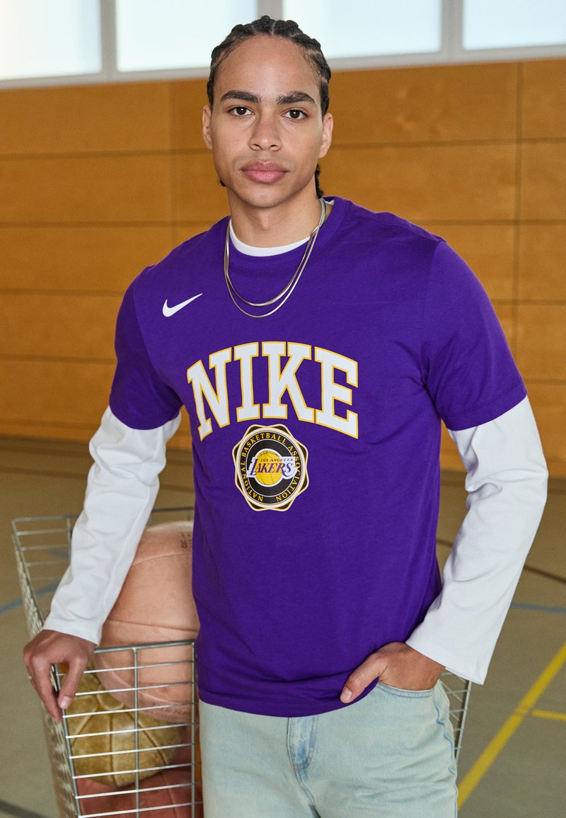 T-shirt violet Nike avec écriture jaune, arborant les logos "NIKE" et "Los Angeles Lakers". Associé à un long t-shirt à manches blanches en dessous.