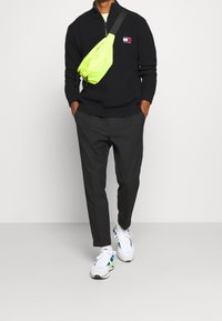 Suéter negro de punto con cremallera, bolsa cruzada verde neón, pantalones grises y zapatillas blancas con acentos azules y verdes.