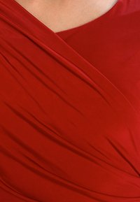 Czerwona sukienka z gładkiego materiału, z drapowanymi fałdami i dekoltem w łódkę. Skupienie na fakturze i jednolitym wzornictwie bez hardware'u i wzorów.
