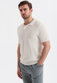 Camisa polo blanca de manga corta con un tejido texturizado, tapeta de dos botones y cuello acanalado, combinada con pantalones grises de rayas.