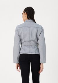 Veste en denim gris clair au waist ajusté, manches longues et col à boutons, avec des coutures surpiquées et un design épuré et minimal.