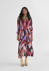 Lunga vestito trasparente con un motivo astratto colorato in rosso, viola e nero. Presenta un colletto, vita con cintura e spacchi laterali. Indossato con stivaletti oro alla caviglia.