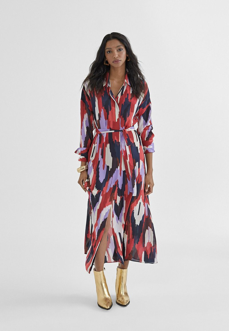 Lunga vestito trasparente con un motivo astratto colorato in rosso, viola e nero. Presenta un colletto, vita con cintura e spacchi laterali. Indossato con stivaletti oro alla caviglia.