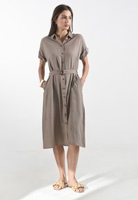 Una donna è in piedi indossando un vestito khaki con cintura, bottoni e sandali intrecciati, con uno sfondo neutro che mette in risalto il suo abbigliamento.