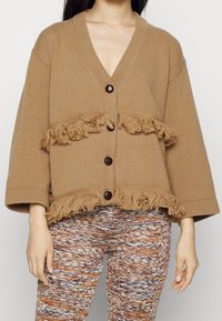 Cardigan marron en tricot avec des manches amples, bordure à franges et trois gros boutons. Porté avec des leggings à motifs.