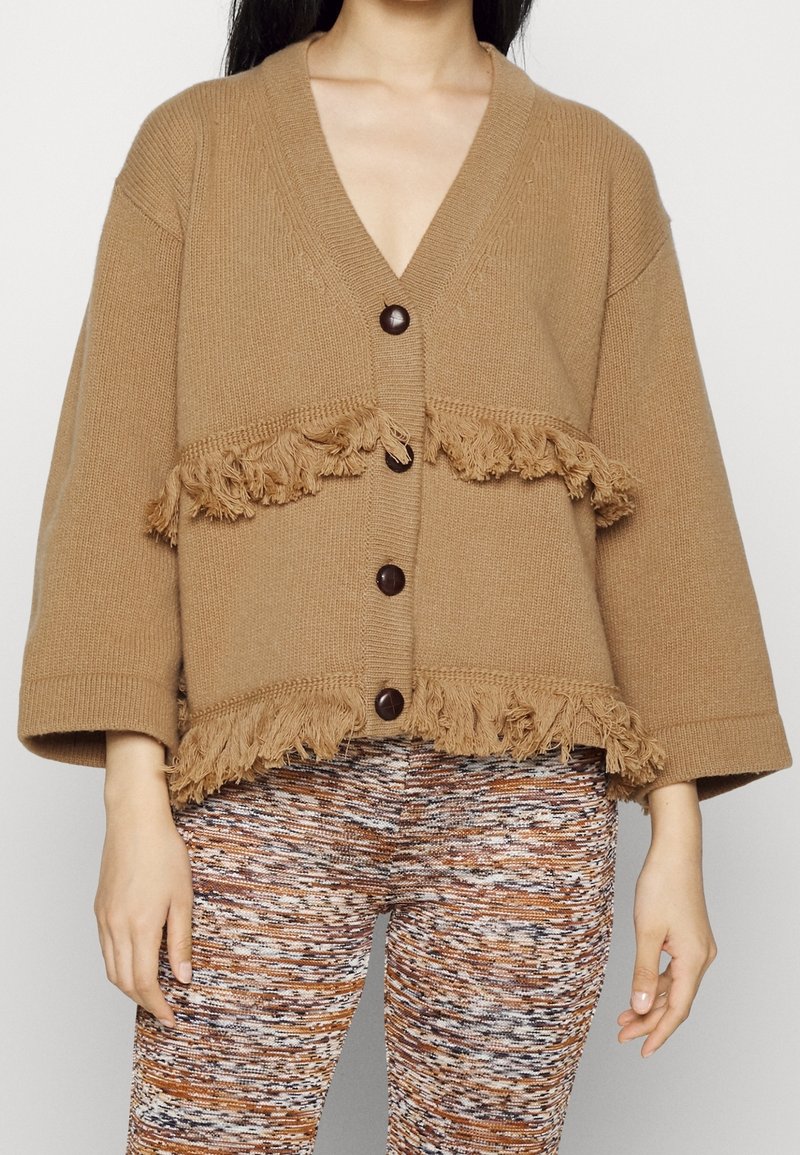 Cardigan marron en tricot avec des manches amples, bordure à franges et trois gros boutons. Porté avec des leggings à motifs.