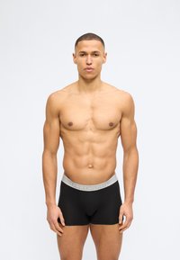 Μυώδης άνδρας με κοντά μαλλιά στέκεται στραμμένος προς τα εμπρός φορώντας μαύρα ανδρικά εσώρουχα Tommy Hilfiger με γκρι ζώνη μέσης σε απλό ανοιχτό φόντο.