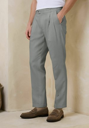 Man wearing light gray pleated trousers with side pockets, beige socks, and brown suede loafers, standing against a beige wall.