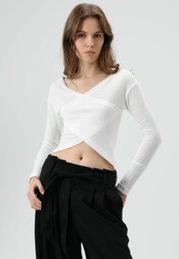 Witte geribbelde top met lange mouwen, een V-hals en een overslagontwerp, gecombineerd met zwarte broek met hoge taille en een ceintuur met knoopdetail.