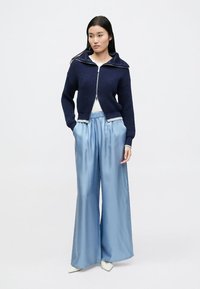 Cardigan bleumarin din tricot cu închidere cu fermoar, model texturat, combinat cu pantaloni largi din satin bleu deschis și top alb. Modelul stă în picioare, drept.