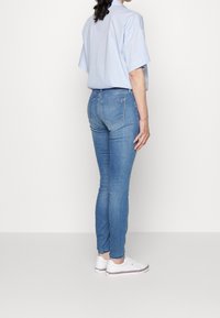 Vaqueros ajustados de color azul claro con una textura suave, diseño de cinco bolsillos y costuras sutiles. Combinados con una camisa de botones suelta y zapatillas blancas.