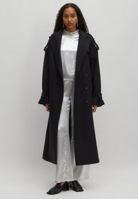 Zwarte lange jas met een op maat gemaakte uitstraling, voorzien van een gesneden kraag, dubbelzijdige knopen en gestructureerde mouwen. Gecombineerd met een glanzende zilveren outfit.