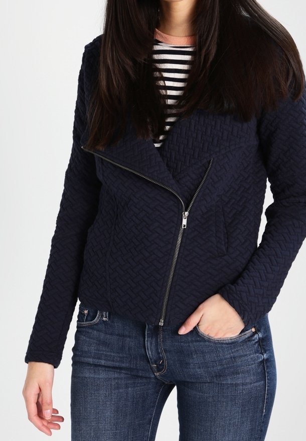 Femme portant une veste matelassée bleu marine avec une fermeture éclair asymétrique sur une chemise rayée noir et blanc et un jean bleu.