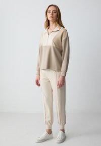 Beige pullover met een rits tot een kwart en een contrasterend borstpaneel, gecombineerd met bijpassende tapered broeken. Beide hebben een zachte textuur.