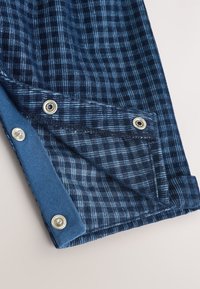 Niebieska tkanina w kratę z metalowymi zatrzaskami, pokazująca częściowo otwartą klapę i miękką podszewkę wewnętrzną.