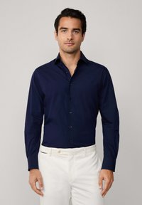 Camisa de manga larga azul marino con cuello clásico y cierre de botones, corte entallado, textura suave, combinada con pantalones blancos.