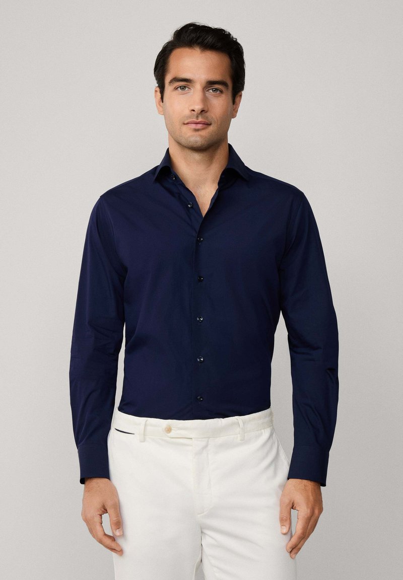 Camisa de manga larga azul marino con cuello clásico y cierre de botones, corte entallado, textura suave, combinada con pantalones blancos.