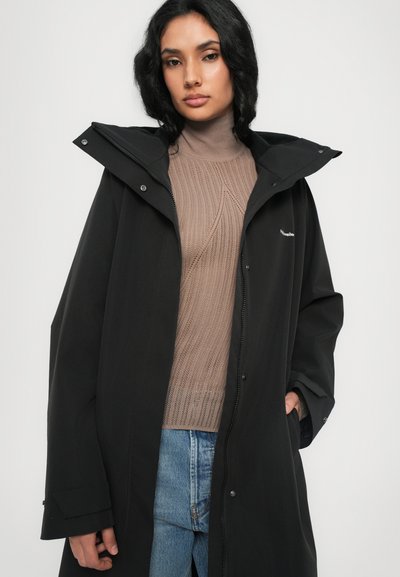 Black waterproof coat with a hood, featuring a front zipper and snap button closures, worn over a light brown ribbed sweater and blue jeans.