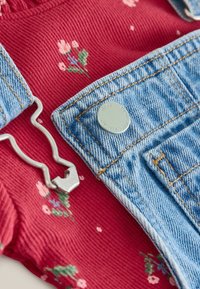 Roter Cordstoff mit floralem Stickmuster, kombiniert mit hellblauen Denim-Trägern, ausgestattet mit einem silbernen Hardware-Haken und einem runden Metallknopf.