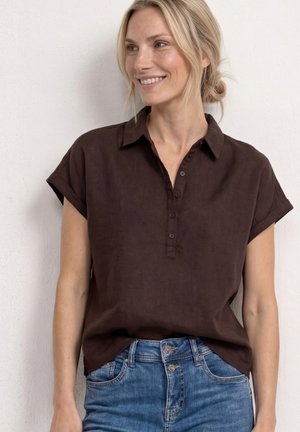 Femme souriante portant une chemise marron foncé à manches courtes boutonnée rentrée dans un jean bleu, debout contre un mur blanc.