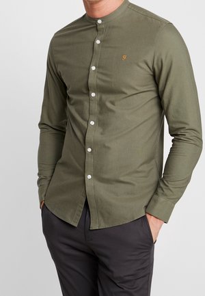 Man in een olijfgroen overhemd met lange mouwen en een mandarinakraag, met witte knopen en donkere broek, handen in de zakken, staand tegen een effen achtergrond.